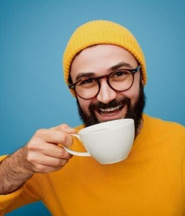 Smiling man wearing a yellow beanie and sweater, holding a white coffee cup against a blue background