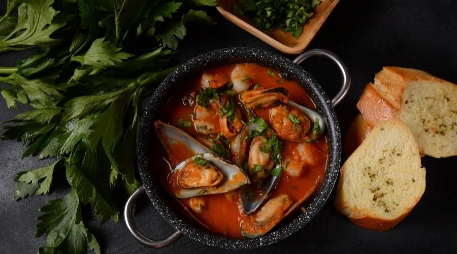 Hearty seafood bouillabaisse soup with mussels and herbs in a black pot, served with garlic bread