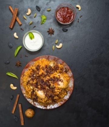 Plate of spiced rice with fried onions and chicken, served with bowls of yogurt and red chutney, surrounded by cinnamon sticks, cashews, and star anise