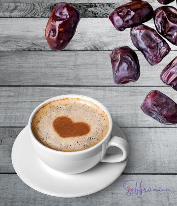 Cup of frothy date seed coffee with heart-shaped cinnamon on top next to fresh dates on wooden table