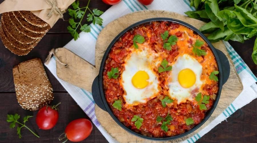 Saffron tomato shakshuka with two sunny-side-up eggs garnished with parsley in a black skillet, surrounded by fresh tomatoes, bread, and basil