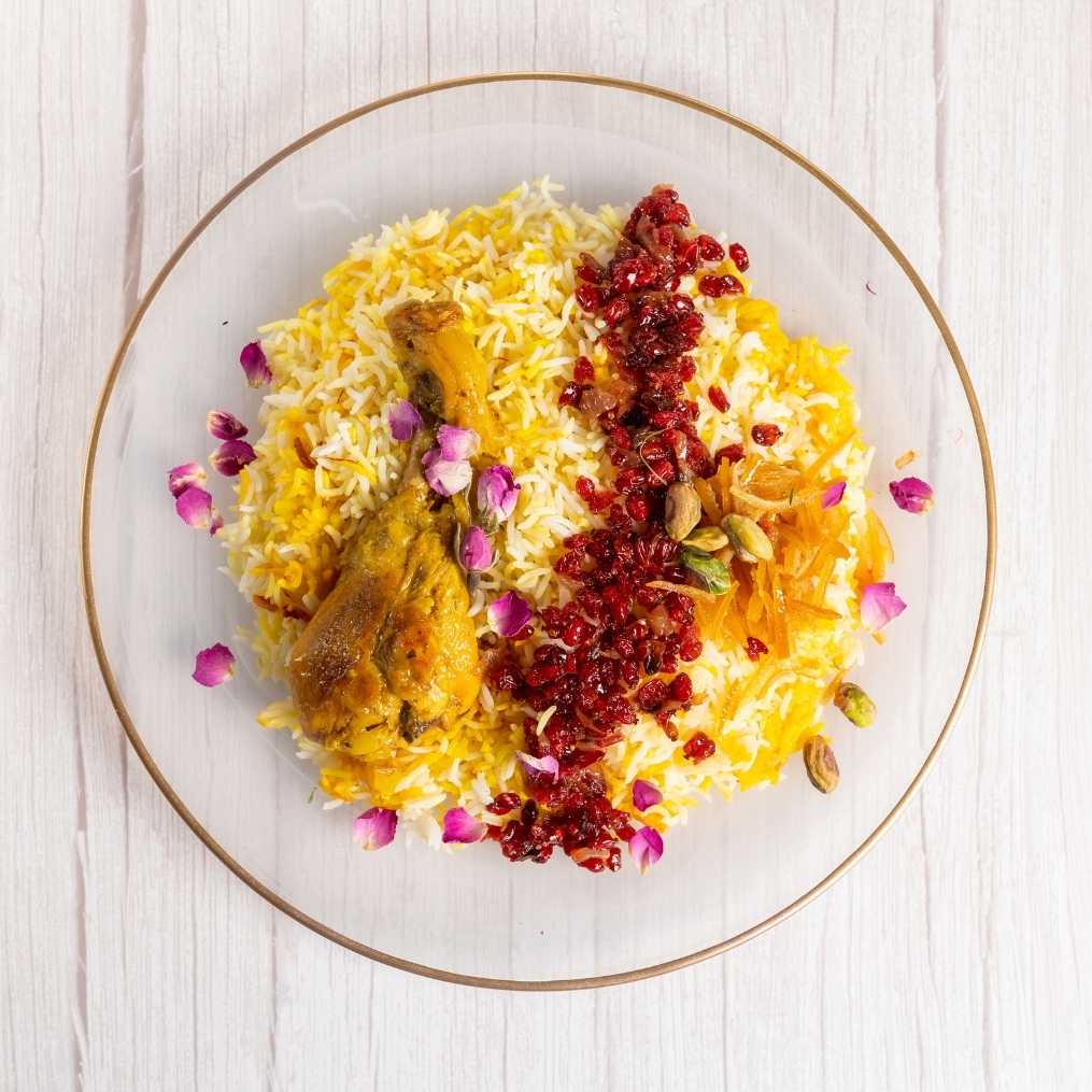 Plate of Zereshk Polo with saffron rice, chicken drumstick, barberries, rose petals, and pistachios on a white wooden table