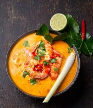 bowl of Thai red curry with shrimp, lime, lemongrass, chili, and kaffir lime leaves on dark wooden table