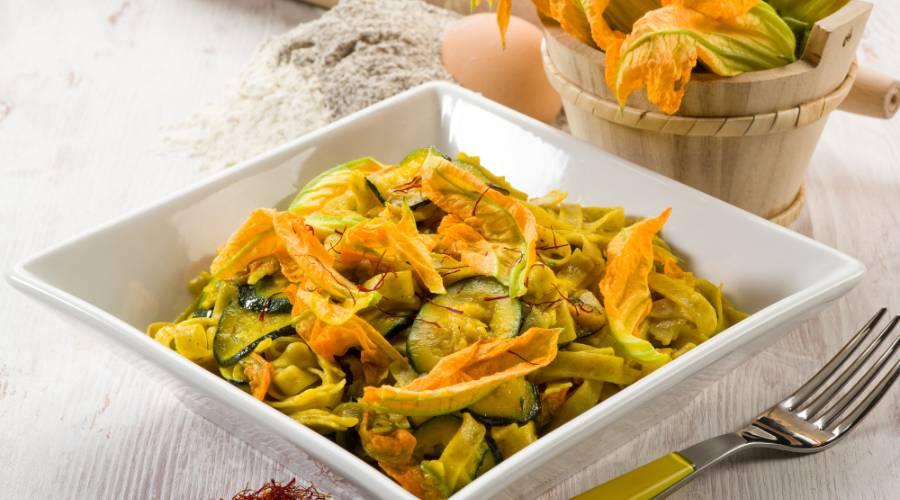 Saffron pasta with zucchini slices and squash blossoms served in a white square bowl on a wooden table