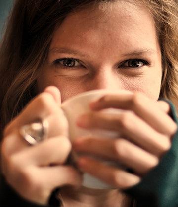 Close-up of woman holding white ceramic cup, smiling with eyes, cozy indoor tea moment